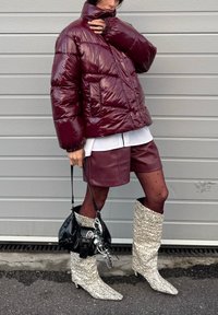 Personne portant une doudoune bordeaux et un short avec des collants noirs à pois, tenant un sac à main noir, chaussée de bottes blanches à motifs montant jusqu'aux genoux.