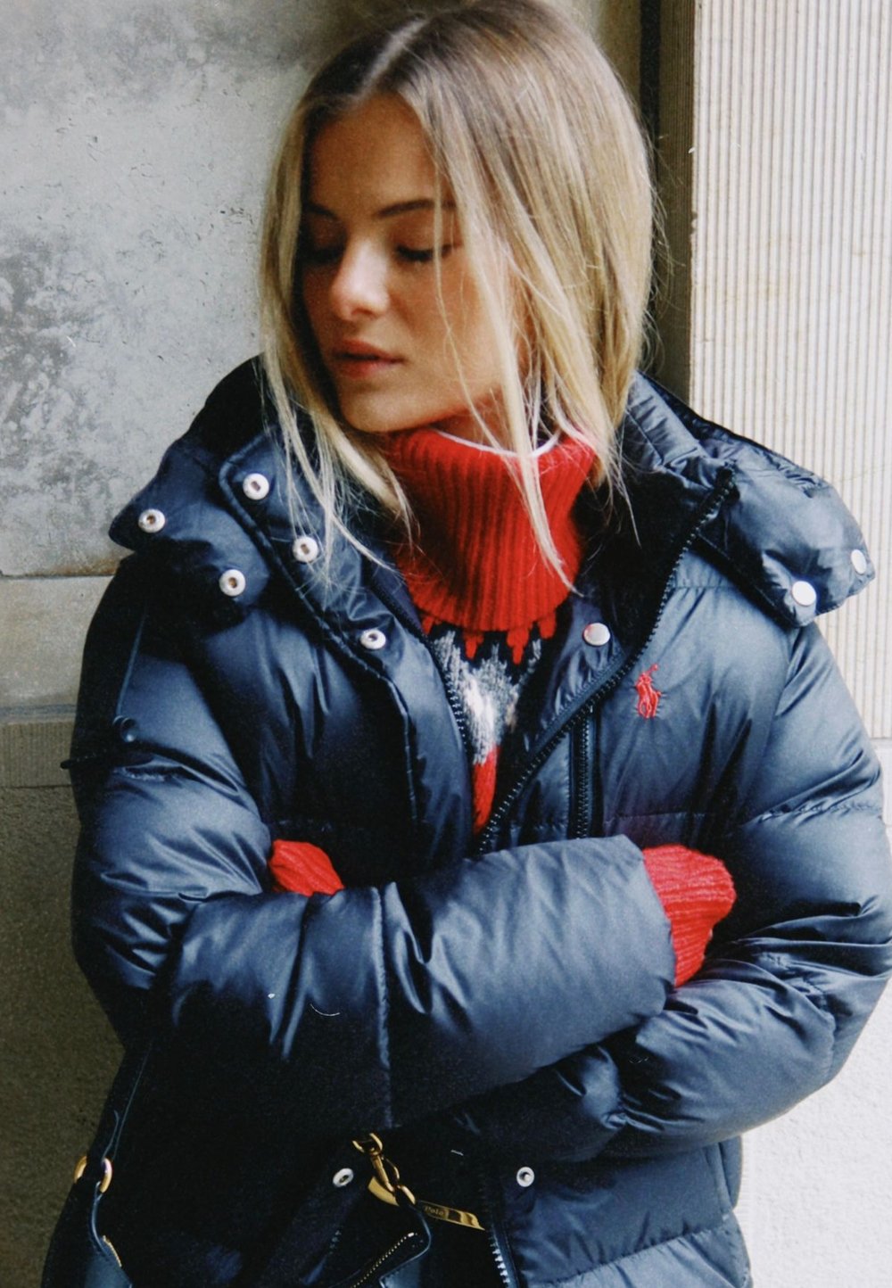 Svart vadderad jacka med blank yta, stor huva och silverfärgade knappar; lager-på-lager med en röd polotröja och mönstrad tröja under.