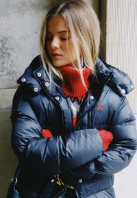 Svart vadderad jacka med blank yta, stor huva och silverfärgade knappar; lager-på-lager med en röd polotröja och mönstrad tröja under.