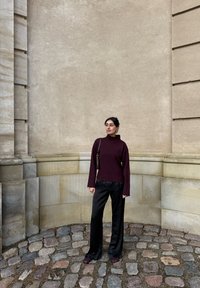 Bourgondische gebreide coltrui, zwarte satijnen broek met wijde pijpen, en bordeauxrode platte schoenen tegen een textuurrijke stenen achtergrond.