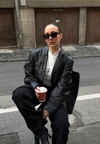 Femme portant une veste en cuir noire et des lunettes de soleil, tenant une tasse de café, assise sur un bollard de rue avec un sac noir tressé derrière elle.