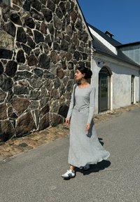 Robe grise à manches longues avec une finition texturée, présentant une taille cintrée et un détail ceinturé. Associée à des baskets noires et blanches, marchant sur le pavé.