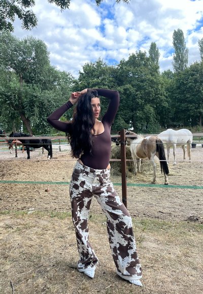 Un bodysuit de manga larga marrón combinado con pantalones de pierna ancha con estampado de vaca en blanco y marrón. La escena incluye caballos y follaje verde en el fondo.