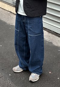 Laisaks lõigatud sinised teksapüksid kontrastõmblustega, millel on suured ees taskud ja lai s�äärekuju, kombineeritud hallide tossudega.