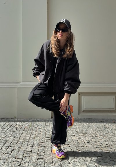 Chaqueta negra oversized, pantalones negros y zapatillas coloridas con acentos en púrpura, amarillo y naranja. Gorra negra y gafas de sol.
