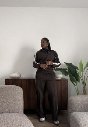 Mujer con chándal marrón oscuro con rayas blancas, de pie en una habitación minimalista entre dos sillones acolchados, sosteniendo un teléfono y mirando de lado.