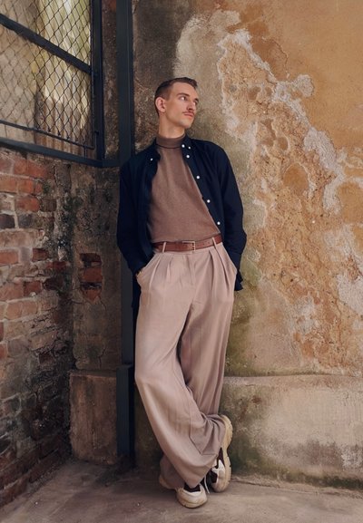 Pantalones de pierna ancha beige, suéter de cuello alto marrón y una camisa de botones oscura. Cinturón de piel y zapatillas blancas. Pared texturizada de fondo.