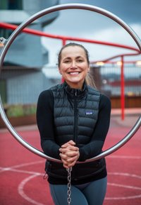 Une femme portant un gilet rembourré noir tient un anneau métallique relié par une chaîne. En arrière-plan, on aperçoit des équipements de terrain de jeu rouges et un ciel gris.