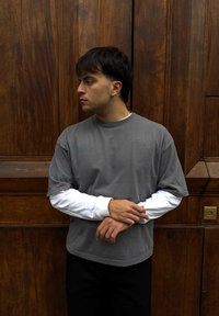 Jeune homme aux cheveux courts et foncés, portant une chemise grise  à manches courtes sur un t-shirt blanc à manches longues, debout devant une porte en bois, regardant vers la gauche.