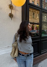 Svītrots garās piedurknes krekls melnbaltā krāsā, gaiši zili džinsi un krēmkrāsas ādas rokassoma. Fona attēlā ir logs un oranža balons.