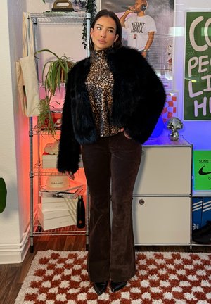 Mujer con chaqueta de piel sintética negra y top de estampado de leopardo, de pie dentro de una habitación junto a una estantería con plantas, zapatos y carteles coloridos en la pared.
