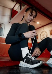 Chaussure de sport noire et blanche avec une semelle épaisse et des lacets larges, attachée par une personne portant une tenue noire à manches longues et des chaussettes blanches.