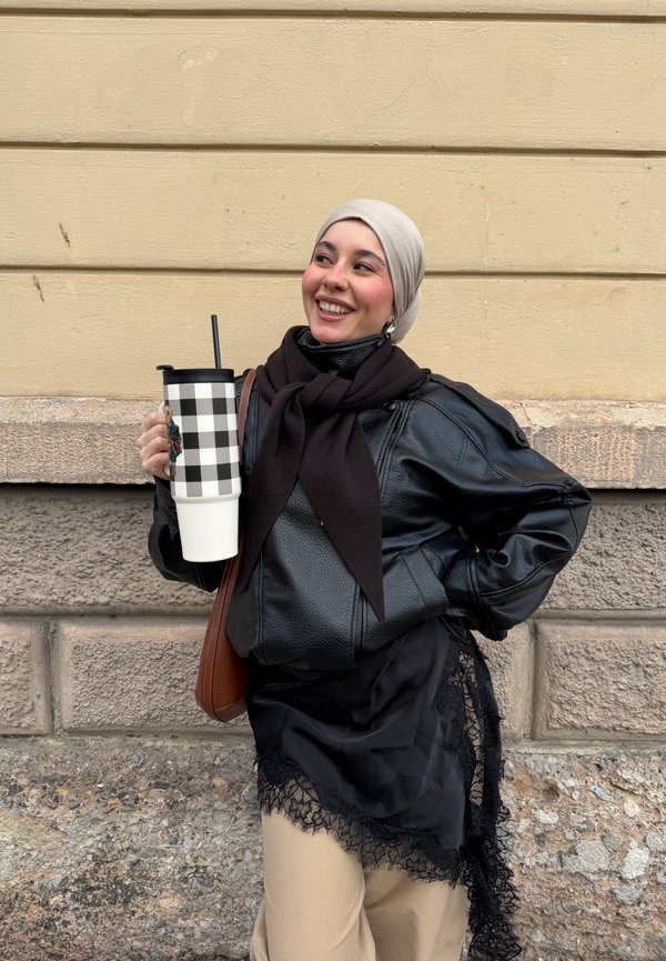 Ung kvinne med beige hodeplagg, svart skinnjakke og skjerf holder en stor rutete termokopp, smilende og stående ved en steinvegg.