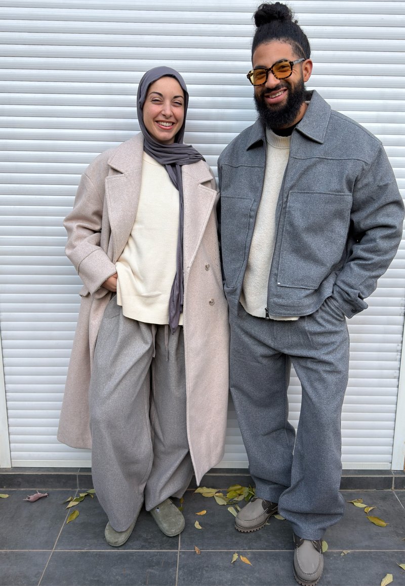 Lächelnder Mann mit Bart und Brille und Frau mit Hijab, beide in gemütlichen, neutral getönten Outfits, stehen im Freien auf gefliestem Boden mit Blättern.