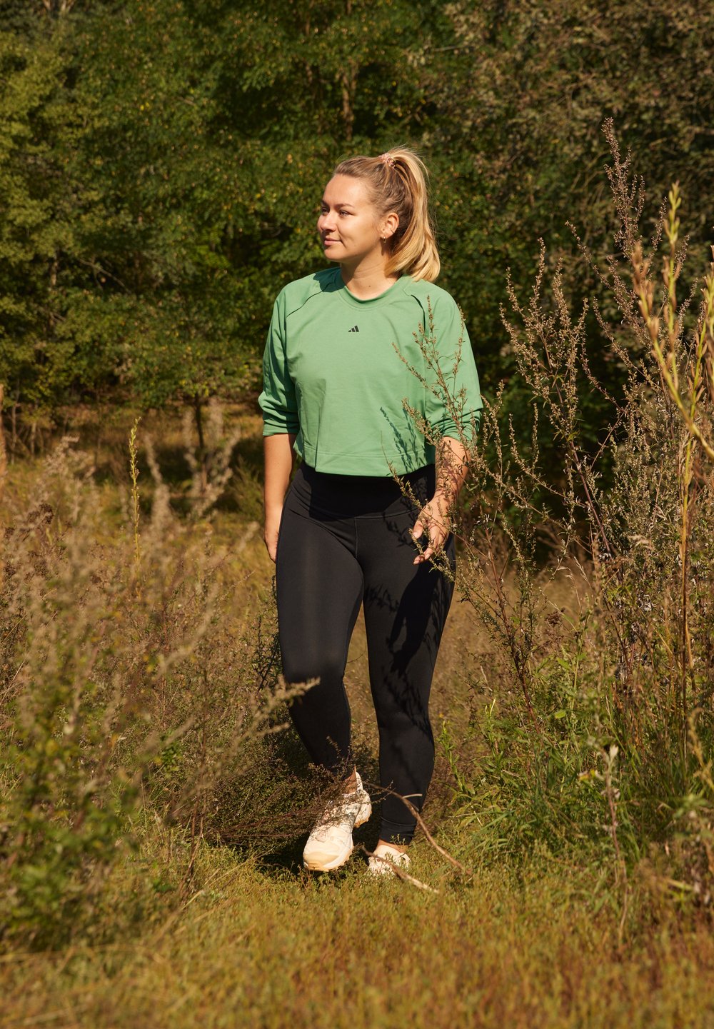 Camisola verde de mangas largas, leggings pretos e sapatilhas de cor clara usadas por uma pessoa a caminhar através de relva alta e arbustos num ambiente natural.
