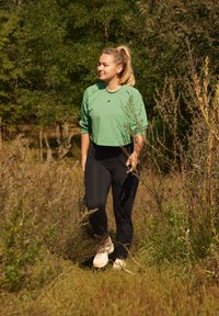 Camisola verde de mangas largas, leggings pretos e sapatilhas de cor clara usadas por uma pessoa a caminhar através de relva alta e arbustos num ambiente natural.