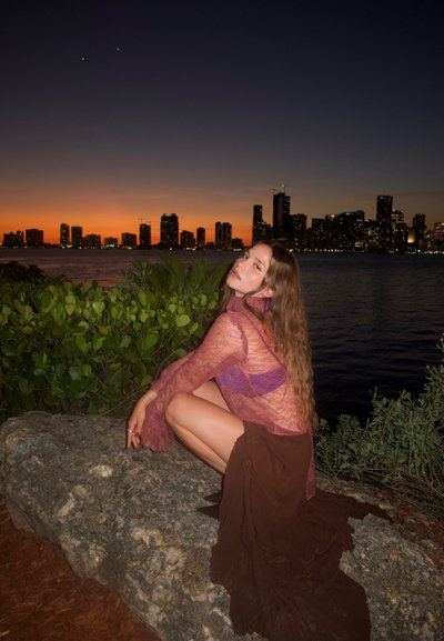 Joven mujer con cabello largo, vestida con una blusa morada translúcida y falda marrón, se agacha sobre una roca junto al agua al atardecer con el horizonte de la ciudad al fondo.