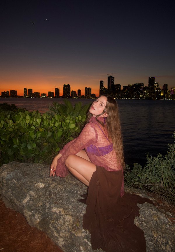 Jeune femme aux longs cheveux portant un haut violet transparent et une jupe marron accroupie sur un rocher près de l'eau au coucher du soleil avec la silhouette de la ville en arrière-plan.