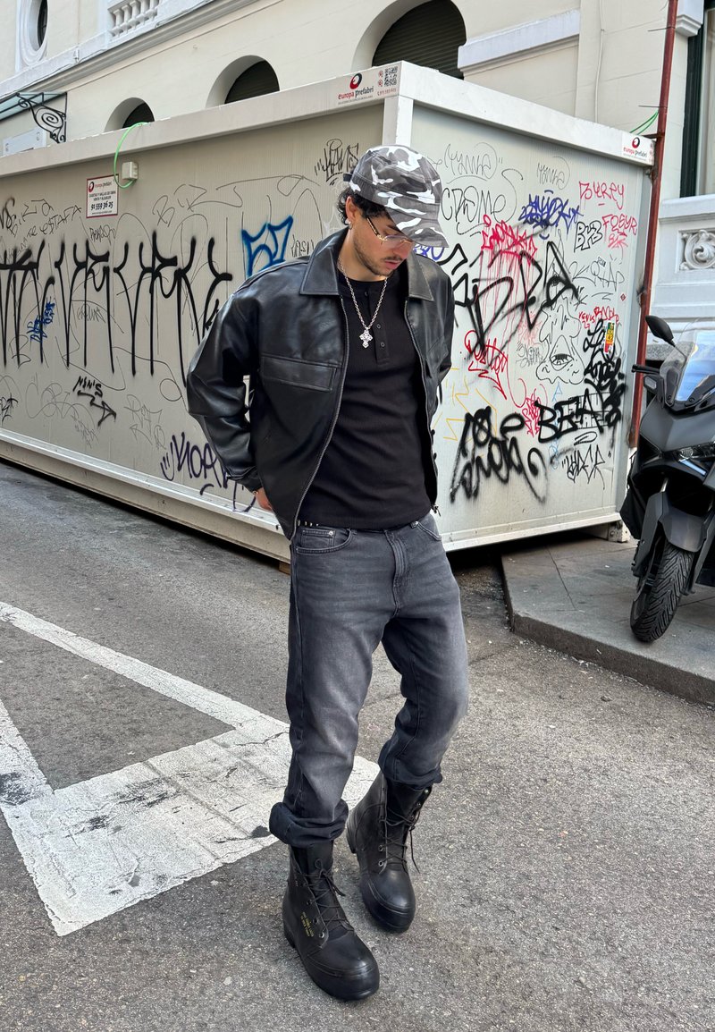 Férfi fekete bőrdzsekiben, terepmintás sapkában, fekete ingben, szürke farmerben és fekete csizmában egy graffiti-szal borított fal közelében áll egy városi utcán.