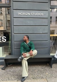 Πράσινο πουλόβερ με πλέξη cable-knit και λευκές λεπτομέρειες, ανοιχτόχρωμο παντελόνι με φαρδιά πόδια και μαύρα αθλητικά παπούτσια. Στο παρασκήνιο υπάρχει ένας γκρίζος τοίχος και μεγάλα παράθυρα.