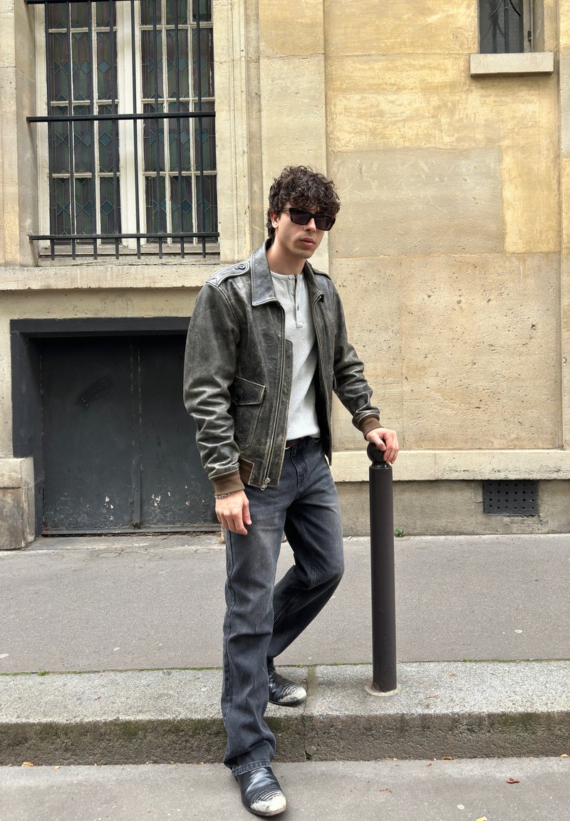 Young man wearing sunglasses and a gray leather jacket leaning on a black post on a city sidewalk beside a beige stone building.