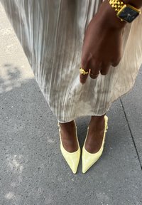Talons aiguilles en cuir verni jaune pâle avec des liens à la cheville, associées à une jupe plissée de couleur claire et des bijoux en or.