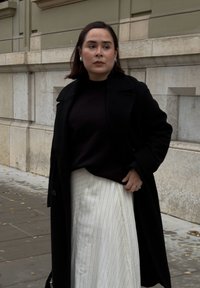 Sort uldfrakke over en mørk sweater, kombineret med et hvidt nederdel med nålestriber. Glatte teksturer og kontrastfarver skaber et lagdelt look.