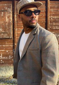 Homme portant un blazer marron à carreaux, une chemise blanche, une casquette à carreaux et des lunettes de soleil foncées, debout à l'extérieur contre un mur en bois usé.