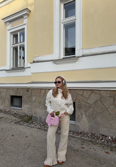 Mujer con blusa de lunares, pantalones beige con volantes, auriculares grises y gafas de sol sostiene una bolsa rosa de punto con flores, de pie junto a una pared de piedra amarilla.