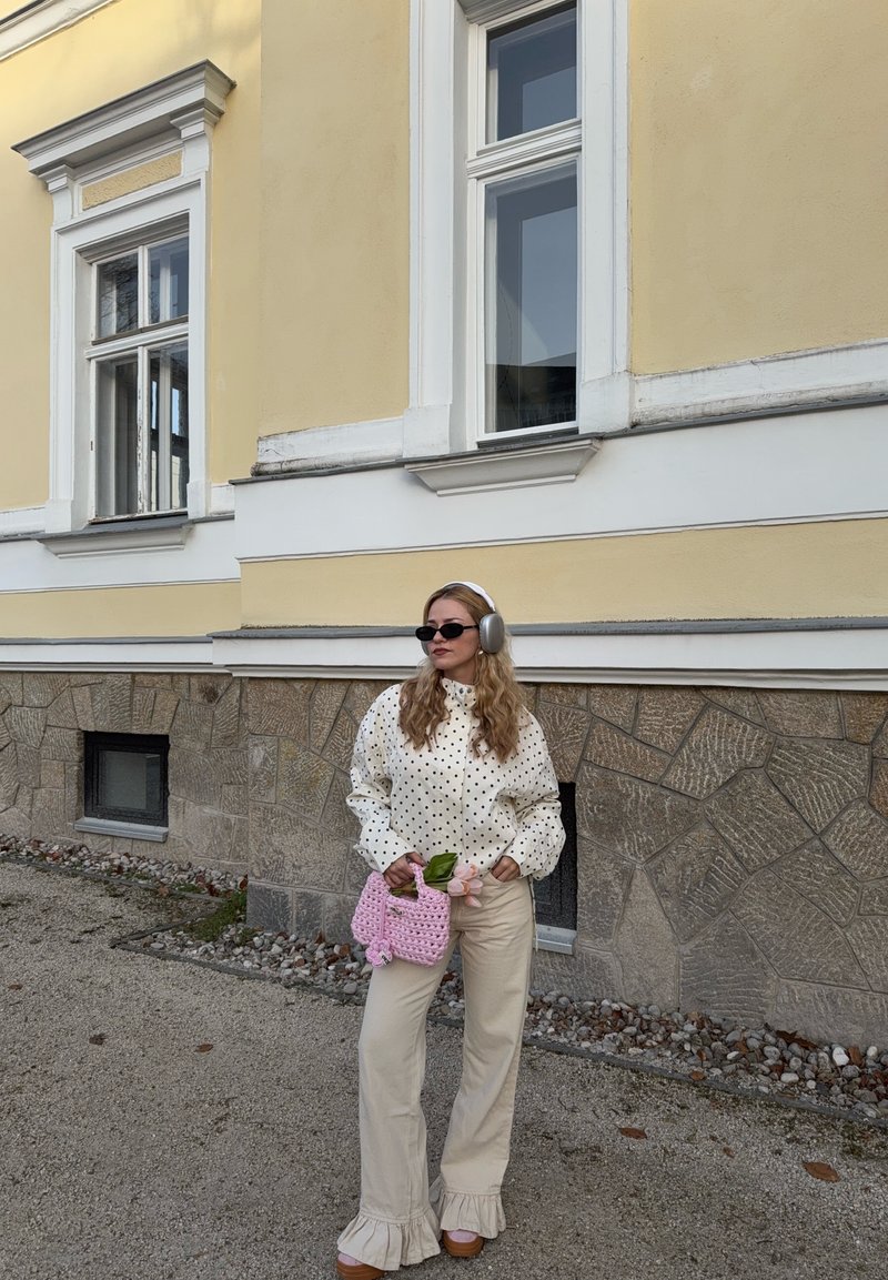 Woman wearing polka dot blouse, beige ruffle pants, gray headphones, and sunglasses holds pink knitted bag with flowers, standing by yellow stone wall.
