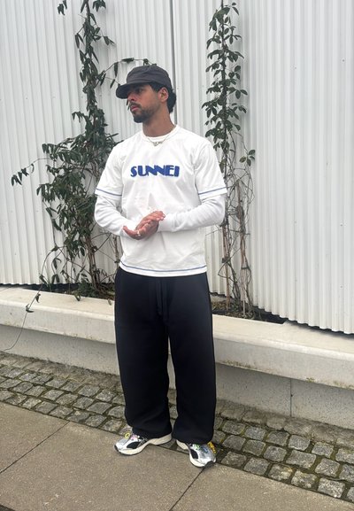 Camiseta blanca con el texto "SUNNEI" en azul marino, combinada con pantalones negros holgados. Los accesorios incluyen una gorra negra y zapatillas multicolores.