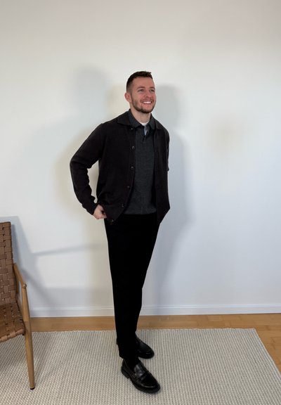 Joven con cárdigan negro, polo oscuro, pantalones negros y zapatos negros, de pie sonriendo contra una pared blanca, cerca de una silla tejida y una alfombra.