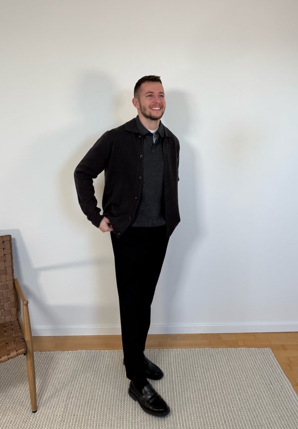 Ung mand i sort cardigan, mørk polo, sorte bukser og sorte sko står smilende mod en hvid væg nær en flettet stol og et tæppe.
