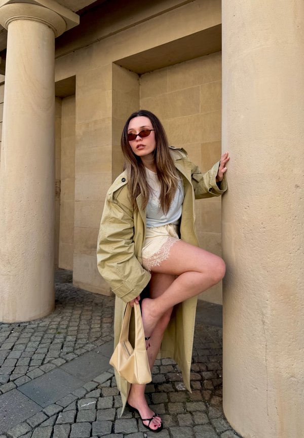 Donna con trench beige, top bianco, shorts in pizzo e occhiali da sole posa tenendo una borsa, appoggiata a una colonna di pietra all'aperto.