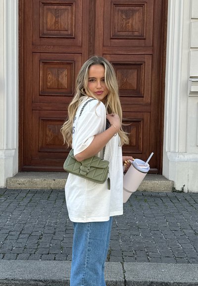 Bolso bandolera acolchado verde con una correa de cadena plateada, camiseta blanca oversize y jeans azules; sosteniendo una gran taza rosa con una pajita.