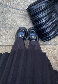 Μαύρη πλισέ φούστα και γυαλιστερά μαύρα παπούτσια με ασημένιο υλικό· μια τσάντα με κυματιστή υφή σε μαύρο χρώμα βρίσκεται δίπλα τους σε μια ποικιλόμορφη επιφάνεια.