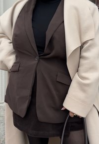 Barna blézer két első zsebbel, fekete felső fölött. Krémszínű, túlméretezett kabát vállakon. Fekete kézitáska látható.