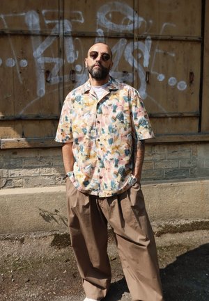 Camisa de manga corta con un patrón floral en colores pastel; pantalones sueltos beige; estilizada con gafas de sol contra un fondo texturizado marrón.