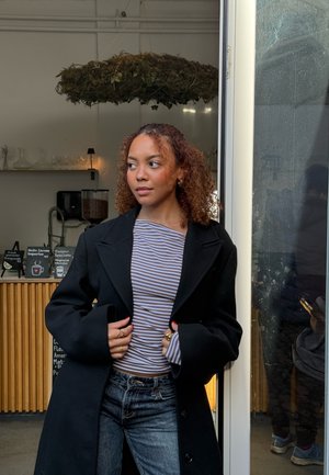 Joven con cabello rizado, vestida con un abrigo negro y una camisa de rayas, de pie en la puerta de un café con un mostrador de madera y un menú de café detrás de ella.