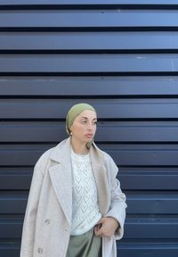 Ανοιχτό μπεζ παλτό, λευκή μπλούζα με σχέδιο καρδιάς, ελαιόχρωμο μαντήλι, σε στάση μπροστά από τοίχο με σκούρες μπλε ρίγες. Καθημερινό ντύσιμο, ουδέτερα χρώματα.