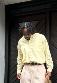 Uomo con dreadlocks che indossa una camicia gialla chiara con bottoni e pantaloni beige, che guarda in basso mentre si trova di fronte a porte in legno scuro.