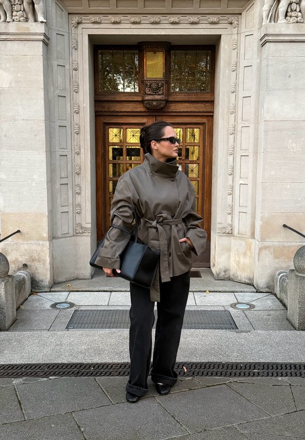 Una mujer con gafas de sol, un abrigo gris con cinturón y pantalones negros está de pie delante de una puerta de madera ornamentada y lleva un bolso negro.