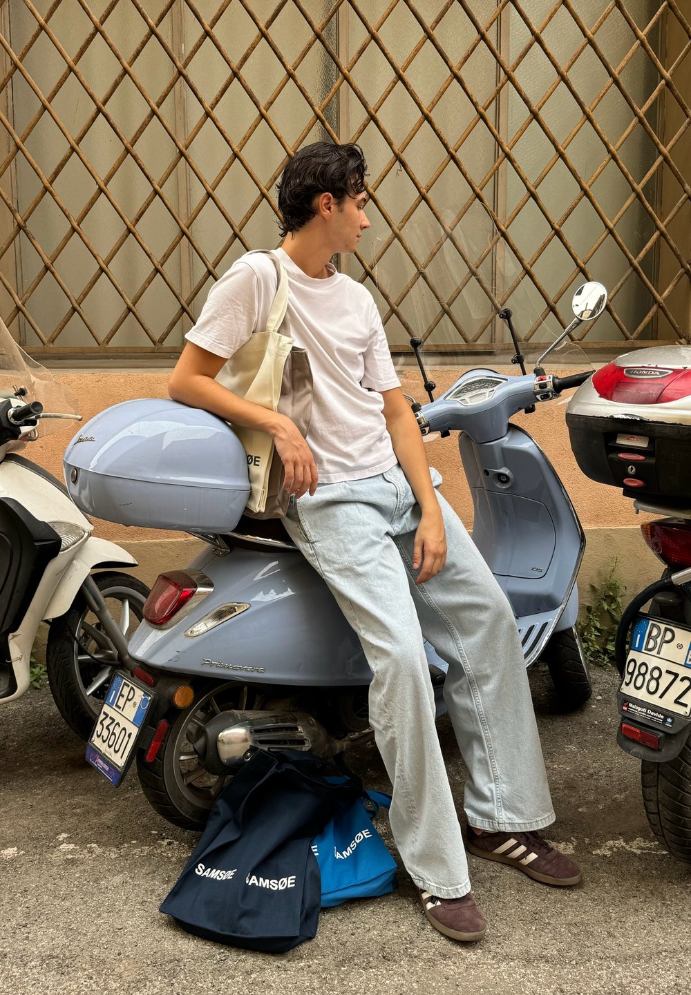 Lys blå scooter med en matchende opbevaringskasse, parkeret ved siden af mørke og lyse tasker, på en tekstureret vej mod en baggrund med geometrisk mønster.