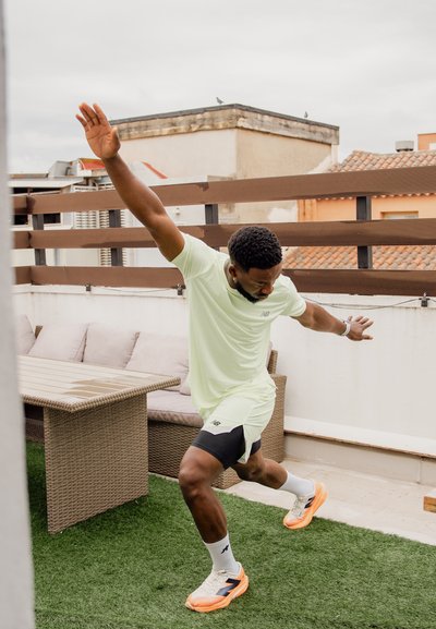 Hombre con ropa deportiva se equilibra sobre césped artificial, con los brazos extendidos, cerca de una mesa de terraza y un sofá de exterior en una azotea.