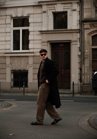 Homme portant un manteau noir et des lunettes de soleil foncées, vêtu d'un pull marron et d'un pantalon beige clair, marchant dans une rue de la ville près d'un bâtiment historique.