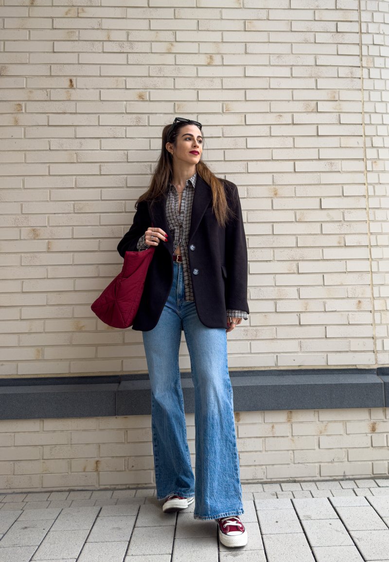 Svart kavaj över en rutig skjorta, utsvängda ljusblå jeans, röd quiltad väska och röda sneakers med vita sulor. Texturerad vägg i bakgrunden.