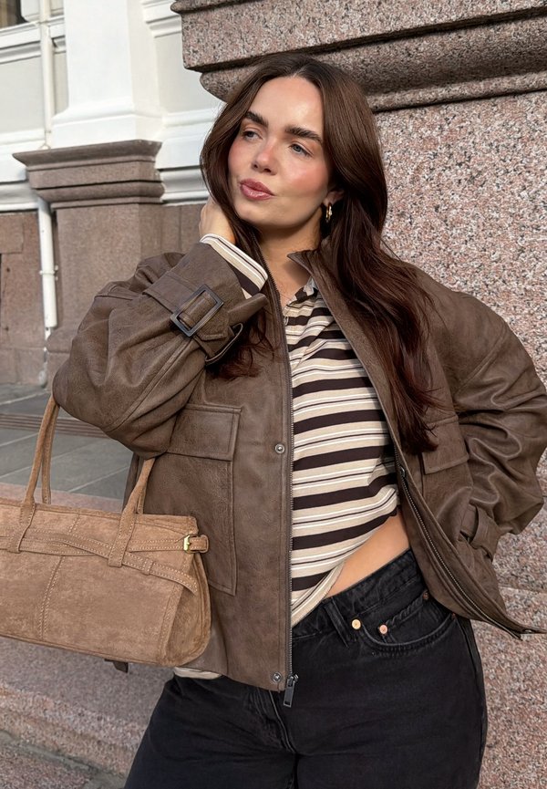 Femme aux longs cheveux bruns portant une veste en daim marron, un crop top rayé, un pantalon noir, et tenant un sac à main en daim beige, posant en extérieur.