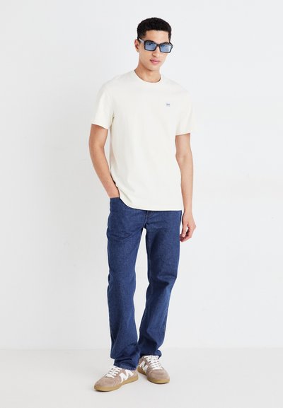 Camiseta de algodón color crema claro con un pequeño logo, combinada con unos jeans de mezclilla azules y zapatillas beige con detalles en blanco y cordones redondos.