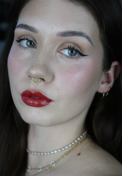 Mujer con ojos verdes, delineador de ojo alado, lápiz labial rojo, anillo dorado en el septum, múltiples pendientes dorados y collares estratificados de perlas y oro, sobre un fondo oscuro.