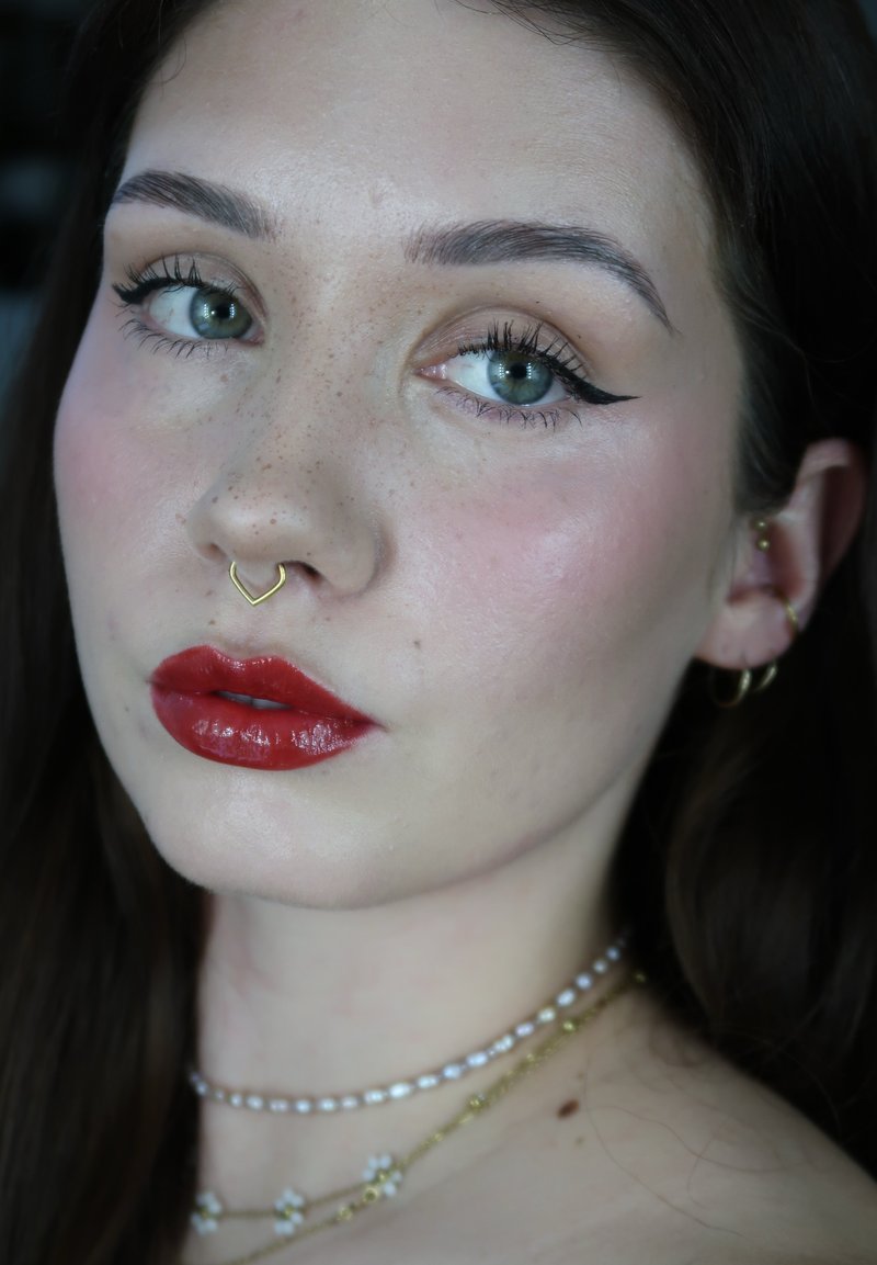 Femme aux yeux verts, eye-liner ailé, rouge à lèvres rouge, anneau de septum en or, plusieurs boucles d'oreilles en or, et colliers superposés en perles et or, sur fond sombre.